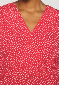 Robe rouge avec un décolleté en V profond et des motifs en forme de cœur blancs. Le tissu semble lisse avec des détails froncés au niveau de la taille.