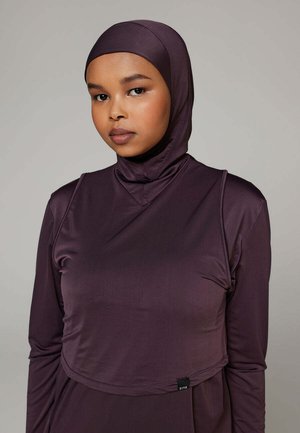 Jonge vrouw die een aansluitende paarse hijab en bijpassende top met lange mouwen draagt, kijkt rustig naar de camera tegen een effen grijze achtergrond.