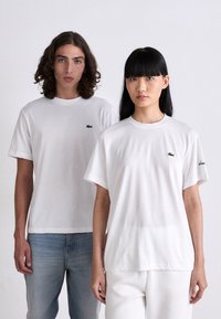 Dos modelos están de pie contra un fondo blanco, vistiendo camisetas blancas de Lacoste; la mujer tiene el cabello negro y el hombre tiene el cabello castaño rizado.