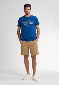 Blå bomulds T-shirt med "Petrol Ind" grafik, parret med beige shorts og hvide sneakers. Casual fit, korte ærmer og rund halsudskæring.