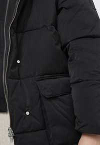 Chaqueta de pluma negra con textura acolchada, que presenta un cierre de cremallera y un bolsillo con solapa en el lateral. Acentos de hardware en color plata.