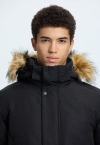 Giacca a vento nera con cappuccio foderato in pelliccia e dettagli beige. I bottoni frontali e una superficie liscia e opaca completano il design.