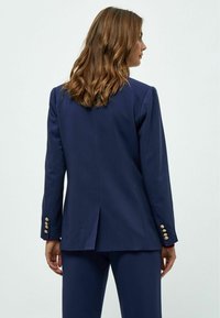 Námořnický modrý blazer s přizpůsobeným střihem, s dvojitým rozparem a zlatými knoflíky na rukávech. Hladká textura látky.
