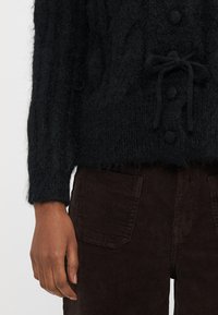 Μαύρο πλεκτό πουλόβερ με σχέδιο καλωδίου, μεγάλα κουμπιά και λεπτομέρεια δεμένης μέσης, σε συνδυασμό με σκ�ούρο καφέ παντελόνι κορντόνι.