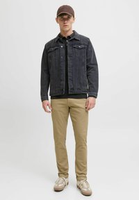 Donker denimjacket met knopenfront, twee borstzakken, gecombineerd met een zwarte poloshirt, beige chinos en beige sneakers met witte accenten.
