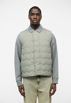 Uomo che indossa un gilet trapuntato grigio chiaro sopra una camicia grigia a maniche lunghe e jeans beige, in piedi contro uno sfondo bianco, rivolto verso l'obiettivo.