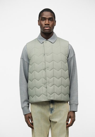 Homme portant une veste matelassée gris clair sur une chemise grise à manches longues et un jean beige, debout devant un fond blanc, faisant face à l'avant.