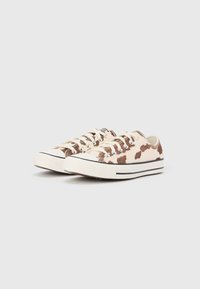 Tenisi cu talpa joasă din pânză, având un design cu imprimeu de vacă în nuanțe bej și maro, tălpici din cauciuc alb și închidere clasică cu șireturi și întărituri metalice.