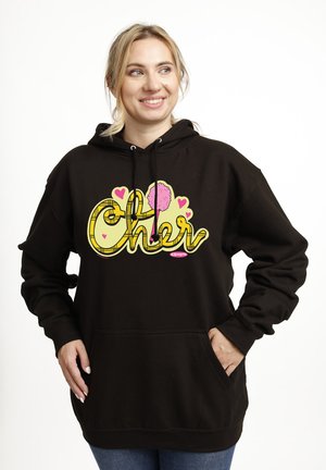 Lächelnde Frau trägt schwarzen Hoodie mit gelbem "Cher"-Schriftzug, rosa Herzen und Pompon-Grafik, Hände in der Fronttasche, heller Hintergrund.