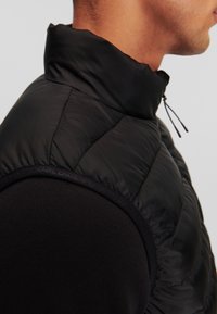 Vesta puffer neagră cu guler înalt și design matlasat, având un material exterior neted și o potrivire strânsă, asociată cu un strat de bază negru cu mânecă lungă.