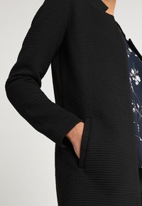 Veste noire texturée au design contemporain, dotée d'un col rond, de manches longues et de poches latérales.
