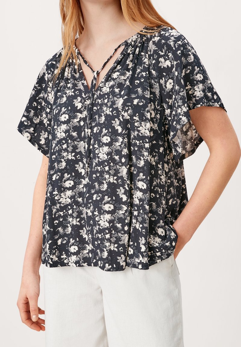 Femme portant un chemisier ample noir à manches courtes fleuri avec un col à lacets, associée à un pantalon blanc, debout avec une main dans la poche.