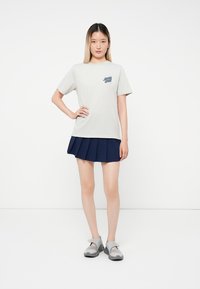 Femeie tânără stând cu mâna pe șold, îmbrăcată cu un tricou deschis la culoare din Santa Cruz, o fustă plisată bleumarin și pantofi de tip Mary Jane gri, pe un fundal alb.