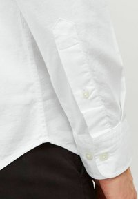 Manche de chemise en coton blanc présentant un poignet aiguisé, fermeture à double bouton et tissu texturé avec un léger éclat. Le bord est taillé avec une finition arrondie.