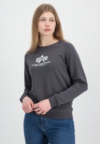 Siva dukserica s okruglim izrezom, rebrastim manžetama i donjim rubom; sadrži bijeli logo "ALPHA INDUSTRIES" na prsima; izrađena od mekog materijala.