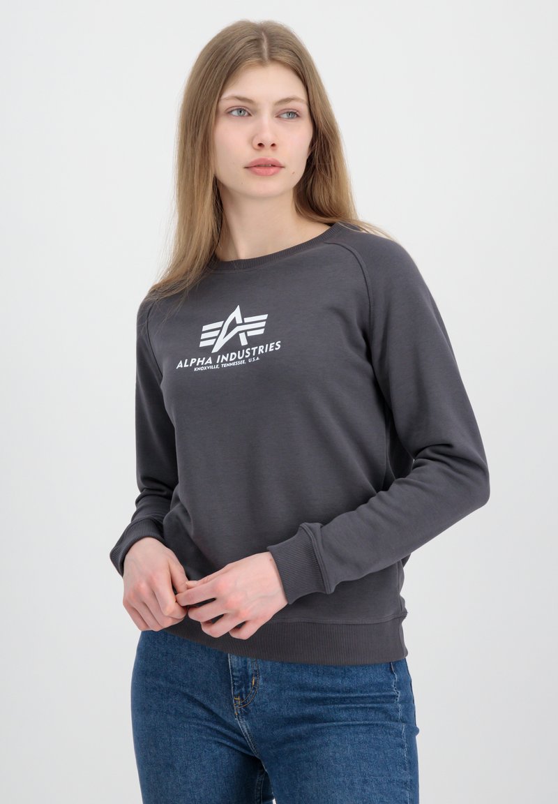 Siva dukserica s okruglim izrezom, rebrastim manžetama i donjim rubom; sadrži bijeli logo "ALPHA INDUSTRIES" na prsima; izrađena od mekog materijala.