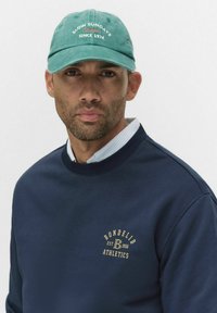 Mies, joka käyttää teal-välistä lippistä, jossa on teksti "Hitaat sunnuntait vuodesta 1974" ja tummansinistä hupparia, jossa on "Bondelid Athletics" -logo rinnassa, valkoinen kauluspaita alla.