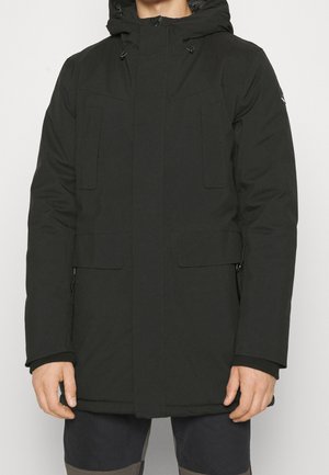 Homme portant un manteau d'hiver noir à capuche avec poches avant et fermeture éclair, se tenant devant un fond blanc uni.