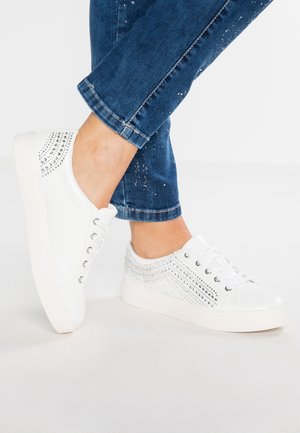 Hvide sneakers med sølvfarvede rhinsten båret med blå denimjeans på krydsede fødder mod en ensfarvet hvid baggrund.