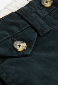 Schwarzer Stoff mit strukturierter Oberfläche, ausgestattet mit einem Knopf mit marmoriertem Muster und einer Klappe mit Stichdetails über dem Knopf.