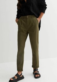 Olive green cotton trousers with a high waist, pleated front, rolled cuffs, and side pockets, paired with black, flat sandals.