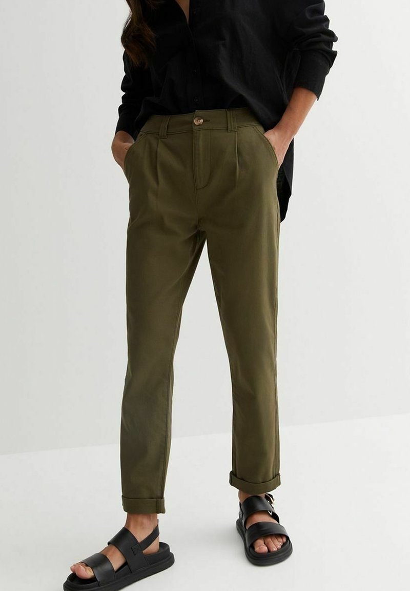 Olive green cotton trousers with a high waist, pleated front, rolled cuffs, and side pockets, paired with black, flat sandals.