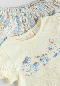Chemise blanche à manches courtes avec broderie florale bleue et marron, associée à un short bleu clair avec un motif floral et une taille froncée.