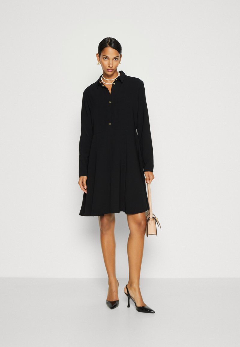 Schwarzes Hemdkleid mit langen Ärmeln, Knopfverschluss, gefälteltem Rock und Kragen. Kombiniert mit schwarzen spitzen Absätzen und einer kleinen beigen Handtasche.