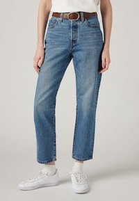 Vaqueros de mezclilla azul con un corte relajado, cintura alta y diseño de pierna recta. Presenta un cinturón marrón y zapatillas blancas con calcetines acanalados.