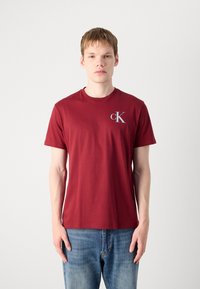 Burgundi pamut póló kerek nyakkivágással, rövid ujjal, az upper bal mellrészen egy kis szürke "cK" logóval. Kék farmernadrággal viselve.