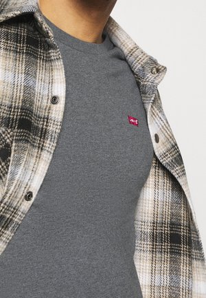 Mężczyzna nosi szarą koszulkę Levi's pod rozpiętą beżowo-czarną koszulą w kratę.