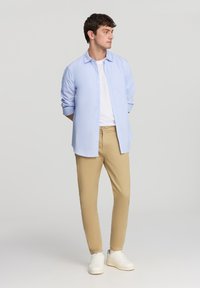 Jasnoniebieska koszula z długim rękawem, biała koszulka pod spodem, beżowe spodnie i białe sneakersy. Model stoi z rękami za plecami na jednolitym tle.