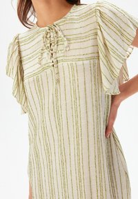 Vaaleanbeige mekko, jossa on vihreät pystysuorat raidat, liehuvat hihat ja nauhakuja, jota kantaa henkilö, joka pitää kättään lantiolla.