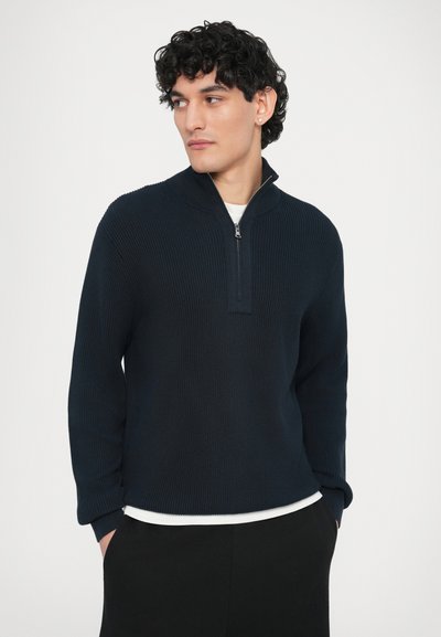 Pull-over côtelé bleu marine avec col zippé au quart, manches longues et coupe décontractée, porté sur un t-shirt à col rond blanc.