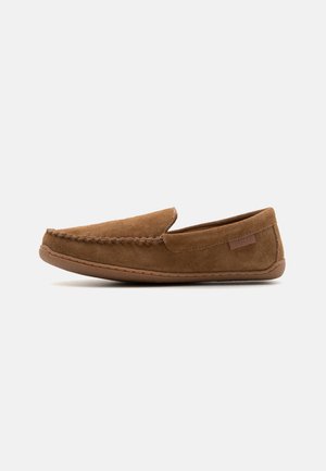 Chaussure en suède marron à enfiler avec un bout arrondi, une couture visible le long du bord et un patch logo subtil sur le côté.