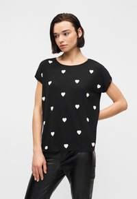 Γυναίκα με μαύρο τοπ με κοντά μανίκια και λευκά σχέδια καρδιών, και μαύρο δερμάτινο παντελόνι, στέκεται με το ένα χέρι πίσω από την πλάτη της.