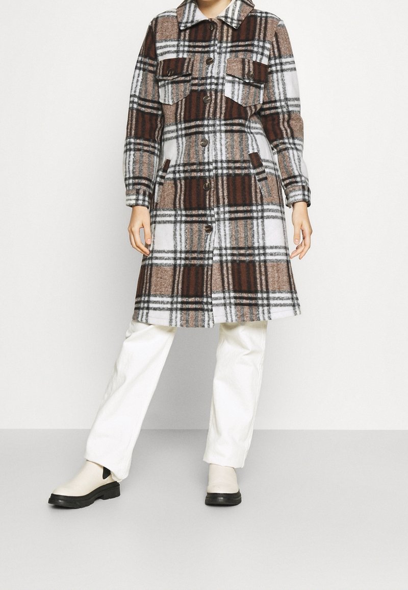 Personne portant un manteau à carreaux marron, blanc et noir jusqu'aux genoux, un pantalon blanc et des bottes claires, debout devant un fond uni.