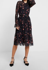 Vestito nero a fiori con maniche lunghe trasparenti, scollo con laccio e orlo a balze; presenta fiori rossi e rosa, vita stretta con cintura.