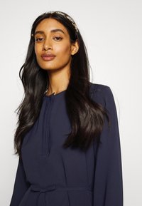 Vestido azul-marinho com um detalhe de pregas subtil na gola, mangas longas e uma cintura atada. Acompanhado por uma faixa de cabelo decorativa.