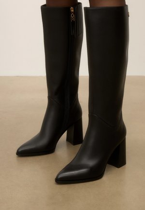 Bottes en cuir noires montant jusque sous le genou, avec des bouts pointus, des talons blocs et des fermetures éclair latérales avec tirettes dorées sur un sol beige.