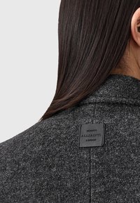 Grijze wollen jas met een gestructureerd oppervlak, voorzien van een zwart leren label met de tekst "ALLSAINTS LONDON", geplaatst bij de halslijn.