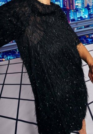 Personne portant une robe noire à texture duveteuse avec des détails scintillants, debout devant un paysage urbain nocturne.