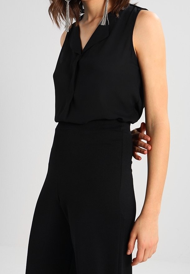 Blouse noire sans manches avec un col et des boutons, associée à un pantalon noir taille haute. Texture lisse, coupe ample.