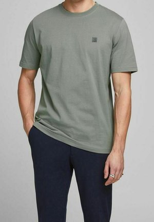 Mand iført en kortærmet olivengrøn t-shirt med et lille logo på brystet og mørkeblå bukser, stående med den ene hånd i lommen.