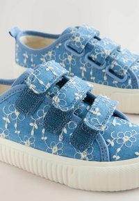 Baskets en toile bleue avec broderies florales blanches, dotées de trois sangles Velcro, d'une semelle en caoutchouc texturée et d'une forme arrondie.