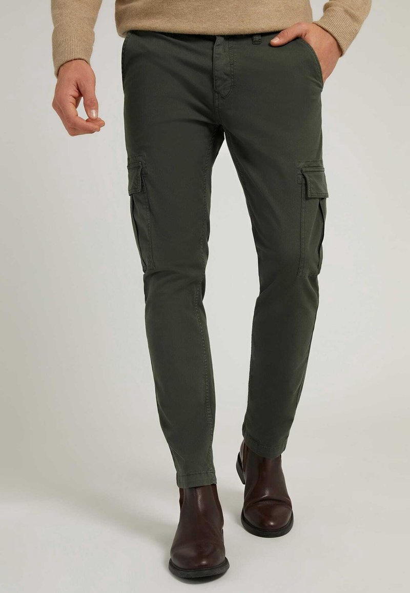 Homme portant un pantalon cargo slim vert olive et des bottines en cuir marron foncé, debout avec une main dans la poche.