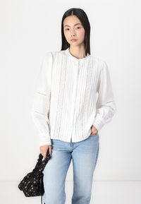 Witte blouse met knoopsluiting, gestructureerde stof, verticale strepen en oogjesdetails. Gepaard met lichtblauwe jeans en een zwarte bloemenhandtas.