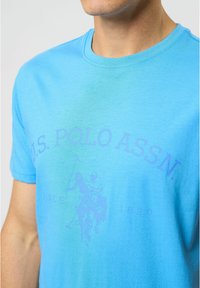 Lyseblå bomulds t-shirt med rund hals, prydet med et falmet lilla grafisk motiv af polo spillere og teksten "U.S. POLO ASSN. SINCE 1890".