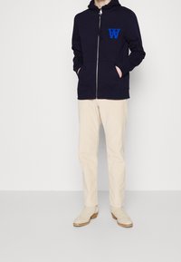Navy zip-up hoodie with front pockets and a blue "W" logo, paired with cream-colored trousers and beige shoes with wooden soles.