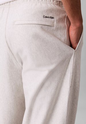 Σκουρό μπεζ παντελόνι Calvin Klein με πίσω τσέπη με πλάγια ραφή και χέρι στην πλευρική τσέπη, φορεμένο από άτομο σε ουδέτερο φόντο.